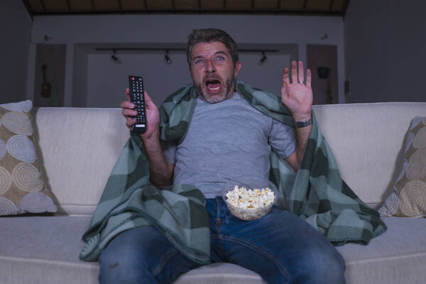 scared and funny man alone at night in living room couch watching horror scary movie in television screaming and eating popcorn covering with blanket