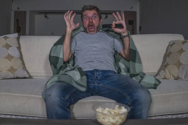 scared and funny man alone at night in living room couch watching horror scary movie in television screaming and eating popcorn covering with blanket