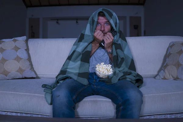 Homme effrayé et drôle seul la nuit dans le canapé salon regarder film d'horreur effrayant ou spectacle effrayant à la télévision manger popcorn couverture avec couverture — Photo