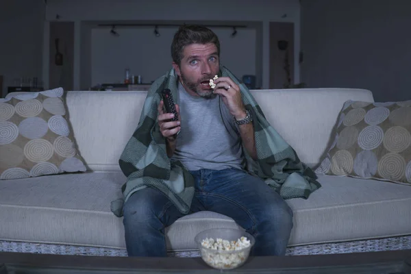 scared and funny man alone at night in living room couch watching horror scary movie or scary show in television eating popcorn covering with blanket