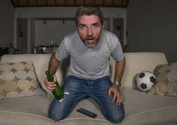 Nervoso ed eccitato tifoso di calcio uomo guardando la partita di calcio in televisione a casa divano sentimento emozione tenendo bottiglia di birra in espressione incredulità — Foto Stock