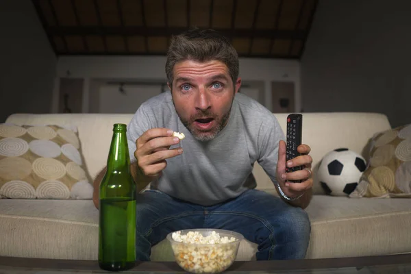 attractive nervous and excited football supporter man watching soccer game on television at home sofa couch in stress and emotion eating popcorn and drinking beer