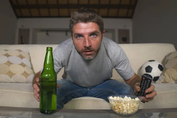 ポップコーンを食べてビールを飲むストレスと感情で自宅のソファソファでテレビでサッカーの試合を見て魅力的な神経質で興奮したサッカーサポーターの男 — ストック写真