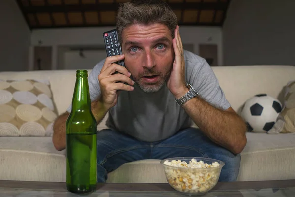 Atraktivní nervózní a vzrušený fotbalista, který sleduje fotbalovou hru v televizi na domácím gauči ve stresu a emoci jíst popkorn a pití piva — Stock fotografie