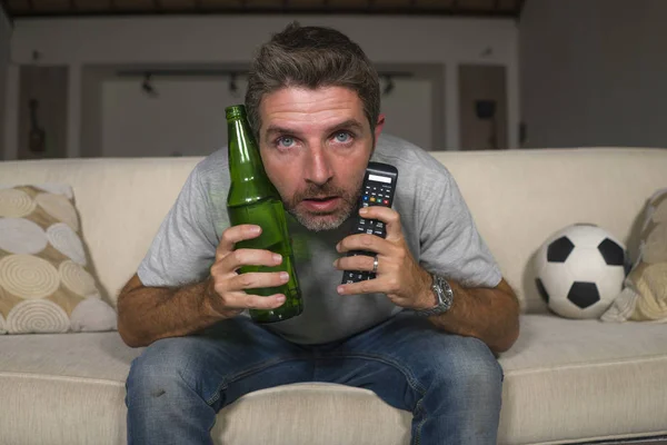 Attraente nervoso ed eccitato tifoso di calcio uomo guardando la partita di calcio in televisione a casa divano divano in stress ed emozione tenendo remoto e bottiglia di birra — Foto Stock