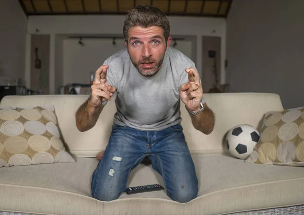 Atractivo nervioso y emocionado partidario del fútbol hombre viendo el partido de fútbol en la televisión en el sofá de la sala de estar sensación intensa emoción y estrés cruzando los dedos — Foto de Stock