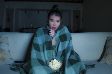  young beautiful scared and frightened Asian Japanese woman watching horror scary movie or thriller eating popcorn in fear face expression eating popcorn on couch clipart