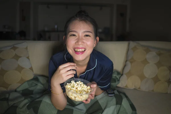 Young beautiful happy and cheerful Asian Korean woman watching TV comedy movie or hilarious show laughing and eating popcorn sitting at home couch with blanket — Stock Photo, Image