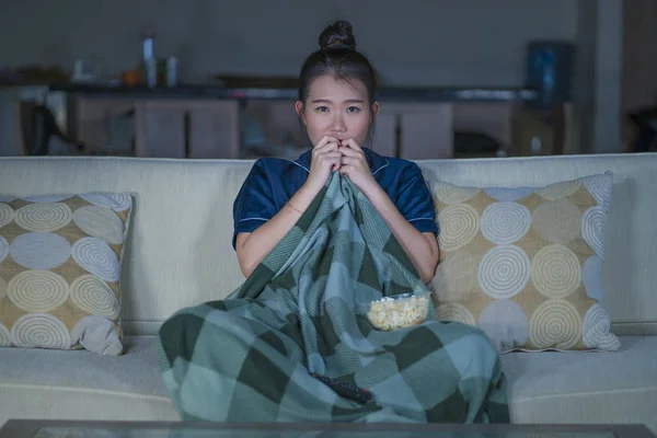 young beautiful scared and frightened Asian Chinese woman watching horror scary movie or thriller eating popcorn in fear face expression eating popcorn on couch