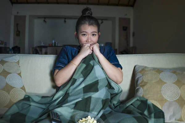 Молодая красивая счастливая и веселая азиатская японка смотрит телевизионный комедийный фильм или смешное шоу смеется и ест попкорн сидя дома на диване с одеялом — стоковое фото