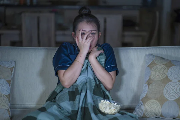 young beautiful scared and frightened Asian Japanese woman watching horror scary movie or thriller eating popcorn in fear face expression eating popcorn on couch