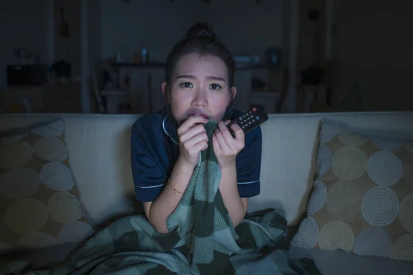 Молодая красивая напуганная и напуганная азиатская японка, смотрящая ужастик страшное кино или триллер, поедающая попкорн в страхе выражение лица, поедая попкорн на диване — стоковое фото