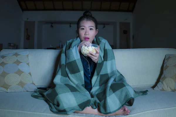 Young beautiful scared and frightened Asian Korean woman watching horror scary movie or thriller eating popcorn in fear face expression eating popcorn on couch — Stock Photo, Image