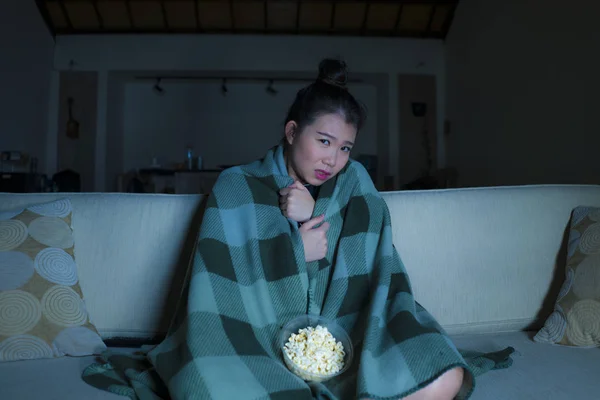 Young beautiful scared and frightened Asian Korean woman watching horror scary movie or thriller eating popcorn in fear face expression eating popcorn on couch — Stock Photo, Image