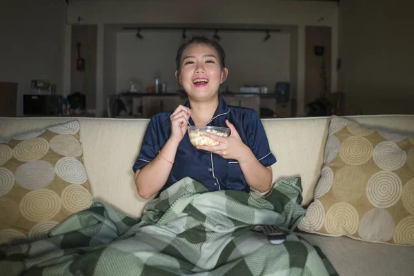 Молодая красивая счастливая и веселая азиатская кореянка, смотрящая телевизионный комедийный фильм или смешное шоу, смеясь и поедая попкорн, сидя дома на диване с одеялом — стоковое фото