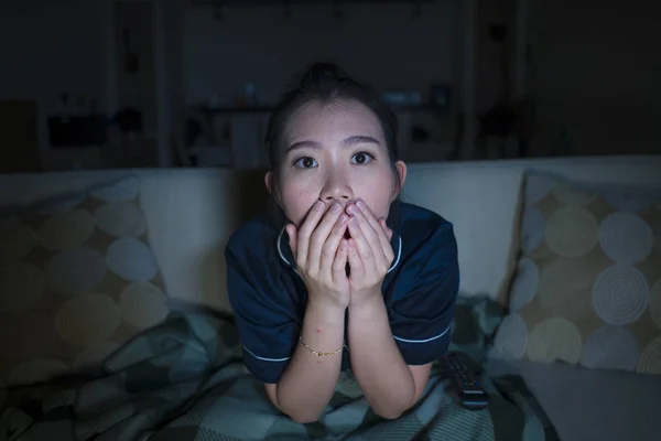 young beautiful scared and frightened Asian Korean woman watching horror scary movie or thriller eating popcorn in fear face expression eating popcorn on couch