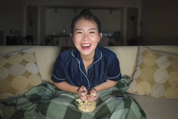 Mladá krásná šťastná a veselá asijská žena, která se dívá na TV komedie nebo legrační show, směje se a žere popcorn, jak sedí doma na gauči s přikrývkou — Stock fotografie