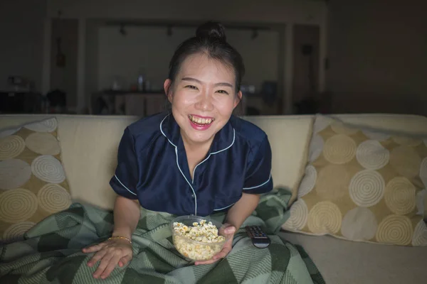 Jeune belle heureuse et joyeuse asiatique femme chinoise regarder la télévision comédie film ou hilarant spectacle rire et manger pop-corn assis à la maison canapé avec couverture — Photo