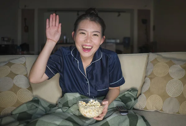 Young beautiful happy and cheerful Asian Korean woman watching TV comedy movie or hilarious show laughing and eating popcorn sitting at home couch with blanket — Stock Photo, Image