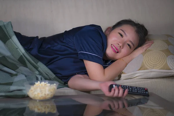 young beautiful happy and relaxed Asian Chinese woman at home living room lying cozy on sofa couch watching TV show episode or romantic comedy movie eating popcorn