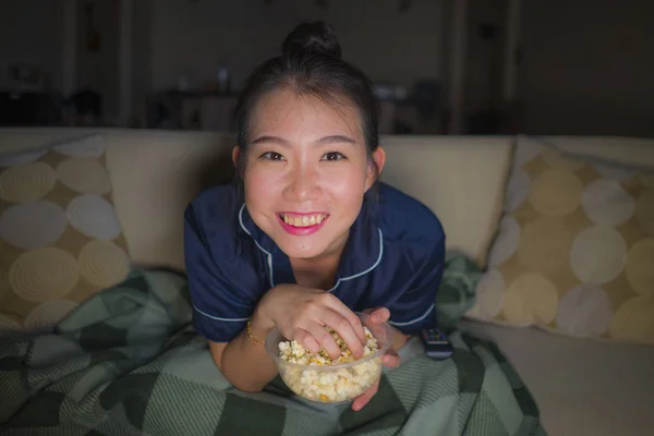 Mladá krásná šťastná a uvolněná asijská žena v obývacím pokoji, kde sedí útulně na pohovce a dívá se na televizní seriál nebo romantický komediální film o jídle popcorn — Stock fotografie