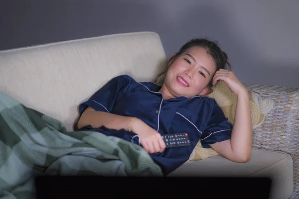 Mladá krásná šťastná a uvolněná asijská žena v obývacím pokoji, kde sedí útulně na pohovce a dívá se na televizní seriál nebo romantický komediální film s úsměvem sladkého — Stock fotografie