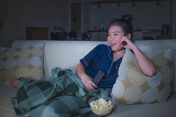 Mladá krásná šťastná a uvolněná Asijská Japonská žena v obývacím pokoji, kde sedí útulně na pohovce a dívá se na televizní seriál nebo romantický komediální film o jídle popcorn — Stock fotografie
