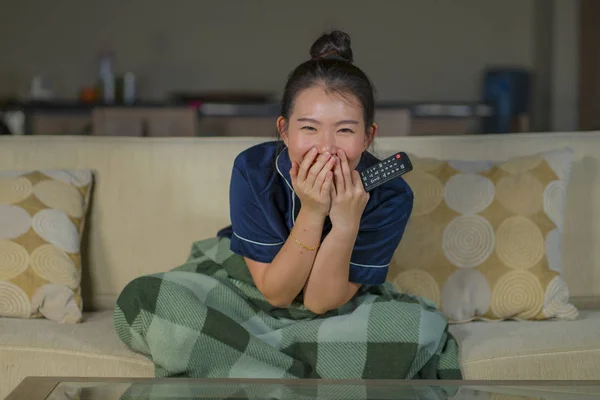Mladá krásná šťastná a veselá asijská žena, která se dívá na TV komedie nebo legrační show, směje se a žere popcorn, jak sedí doma na gauči s přikrývkou — Stock fotografie