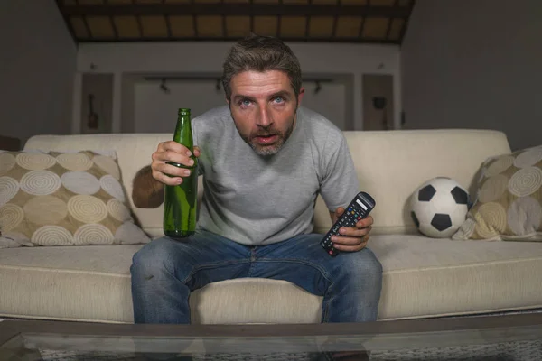 Attraente nervoso ed eccitato tifoso di calcio uomo guardando la partita di calcio in televisione a casa divano divano in stress ed emozione tenendo remoto e bottiglia di birra — Foto Stock