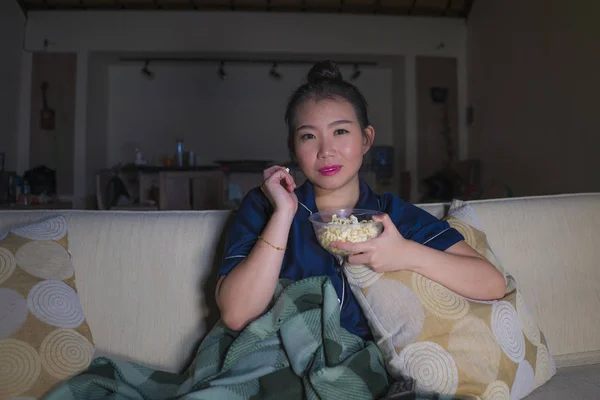 young beautiful happy and relaxed Asian Chinese woman at home living room sitting cozy on sofa couch watching TV show episode or romantic comedy movie eating popcorn