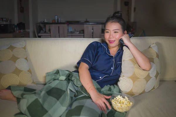 young beautiful happy and relaxed Asian Chinese woman at home living room sitting cozy on sofa couch watching TV show episode or romantic comedy movie eating popcorn