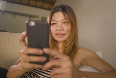 genç çekici ve rahat 30s Asya Endonezya kadın ev oturma odası kanepede cep telefonu ile internet sosyal medya veya online dating uygulaması kullanarak rahat yalan 