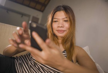 genç çekici ve rahat 30s Asya Endonezya kadın ev oturma odası kanepede cep telefonu ile internet sosyal medya veya online dating uygulaması kullanarak rahat yalan 