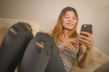 genç çekici ve rahat 30s Asya Endonezya kadın ev oturma odası kanepede cep telefonu ile internet sosyal medya veya online dating uygulaması kullanarak rahat yalan 