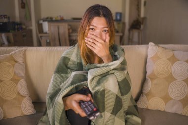 Genç korkmuş veya sürpriz Asya Endonezya kadın izlerken televizyon korkutucu korku film battaniye ile kaplı oturma odasında kanepe kanepede TV uzaktan