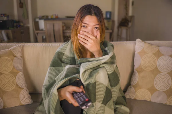 Mladý vystrašený nebo překvapený asijský indonéský ženský, který se dívá na televizi strašidelný horor, pokrytý přikrývkou přidrženou TV dálkový na pohovce v obýváku — Stock fotografie