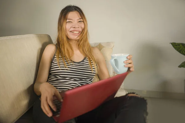 Unga attraktiva och vackra asiatiska indonesiska kvinna hemma arbetar avslappnad och glad med bärbar dator skrattar använda sociala medier sitter på vardagsrummet — Stockfoto