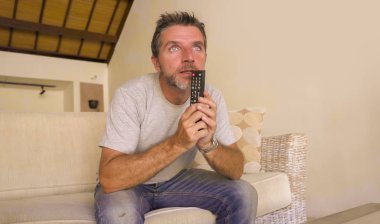 young attractive nervous and excited man sitting at home living room couch holding TV remote watching football game or suspense movie in intense face expression clipart