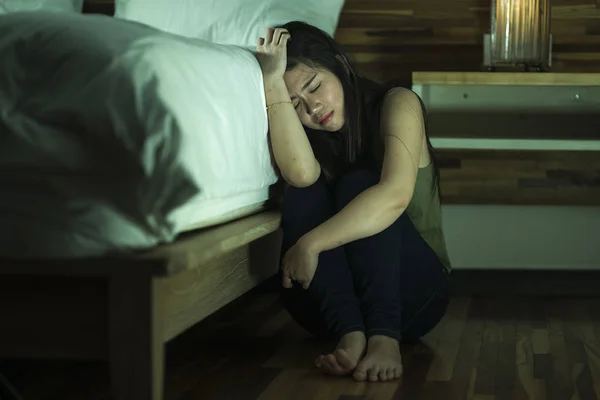 Молодая депрессивная и грустная азиатская японка, сидящая на полу в спальне рядом с кроватью, страдает депрессией и тревожным кризисом — стоковое фото