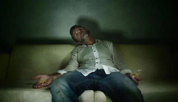 Dramatisches Porträt eines jungen attraktiven depressiven und tablettensüchtigen Mannes, der eine Flasche mit Antidepressiva in der Hand auf der Couch im Drogensuchtkonzept sitzt — Stockfoto