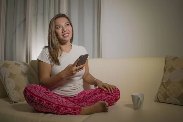 Genç çekici mutlu ve rahat Asya Endonezyalı kadın gülümseyerek cep telefonu internet sosyal medya veya flört uygulaması kullanarak oturma odası kanepede ev keyfi — Stok fotoğraf