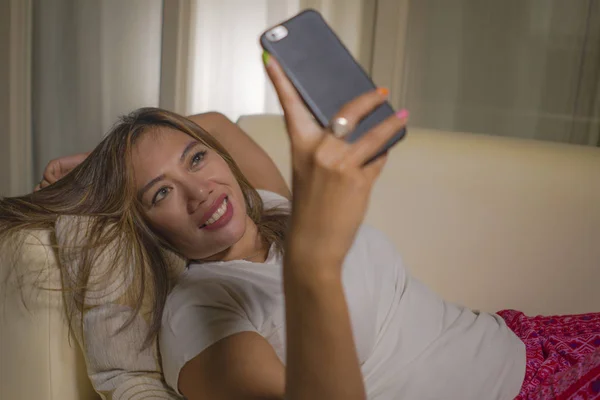 Молодая привлекательная счастливая и расслабленная студентка наслаждается домашним диваном в гостиной с помощью социальных сетей или приложения для знакомств на мобильном телефоне, улыбаясь — стоковое фото