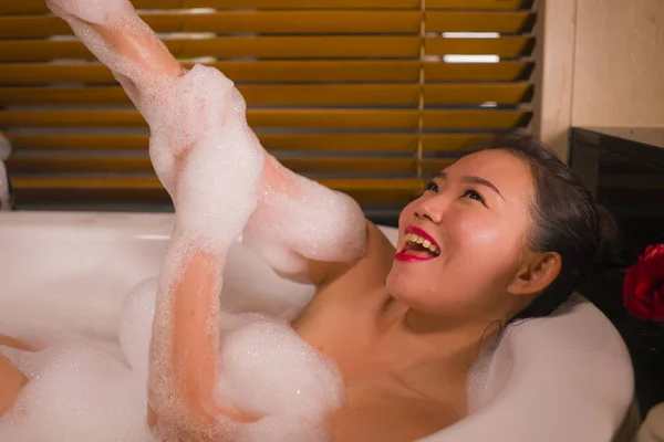 Молодая красивая счастливая и жизнерадостная азиатская кореянка купается дома или в роскошной ванной комнате отеля, купаясь в пенной ванне, наслаждаясь расслабленной улыбкой — стоковое фото