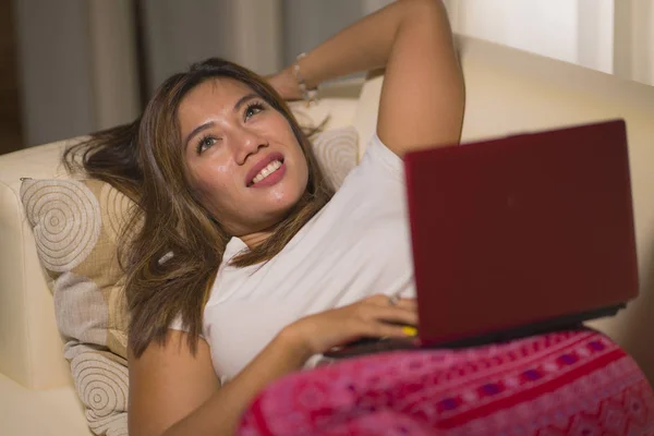 Молодая красивая и естественная азиатская индонезийская женщина в пижаме брюки расслаблены на диване сети с помощью ноутбука покупки в Интернете весело в мечтательный выражение лица — стоковое фото