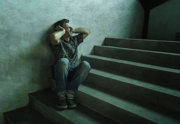 Dramatisches Porträt eines jungen depressiven und paranoiden Mannes, der im Freien auf der Treppe einer dunklen Grunge-Straße an der Ecke sitzt und sich krank fühlt und Depressionen und Angstzustände erleidet — Stockfoto