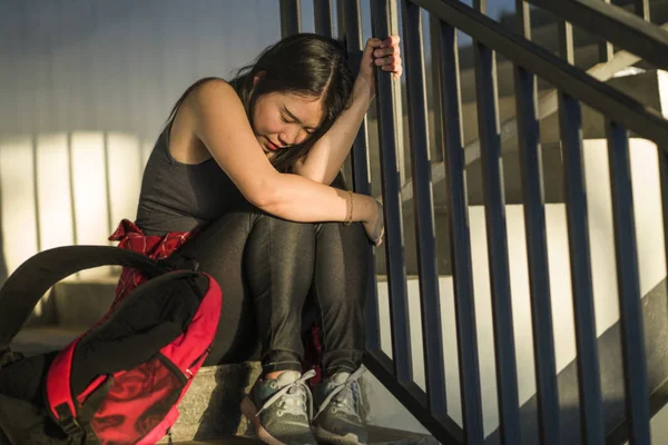 Retrato dramático de una estudiante universitaria asiática acosada. Joven deprimida y triste chica china sentada sola en la escalera del campus sufriendo intimidación y acoso — Foto de Stock