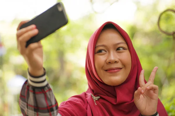 Estilo de vida estudantil muçulmano - jovem bela menina estudante asiática feliz e alegre Indonésia vestindo Islã tradicional hijab cabeça cachecol tomando selfie com telefone celular — Fotografia de Stock