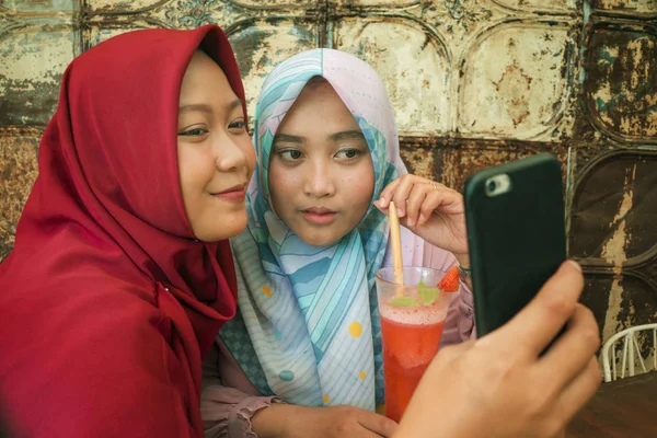 Meninas indonésias felizes e bonitas no tradicional lenço de cabeça hijab islâmico tirando selfie juntos se divertindo no café em amizade e conceito de mídia social — Fotografia de Stock