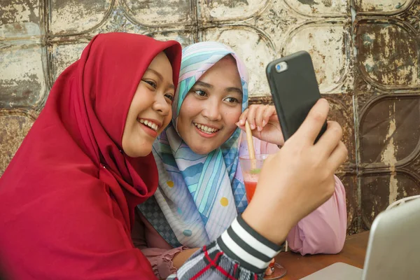 Meninas indonésias felizes e bonitas no tradicional lenço de cabeça hijab islâmico tirando selfie juntos se divertindo no café em amizade e conceito de mídia social — Fotografia de Stock