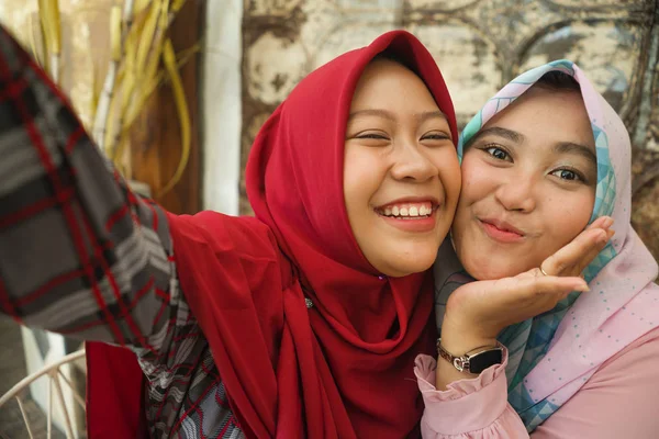 Meninas indonésias felizes e bonitas no tradicional lenço de cabeça hijab islâmico tirando selfie juntos se divertindo no café em amizade e conceito de mídia social — Fotografia de Stock